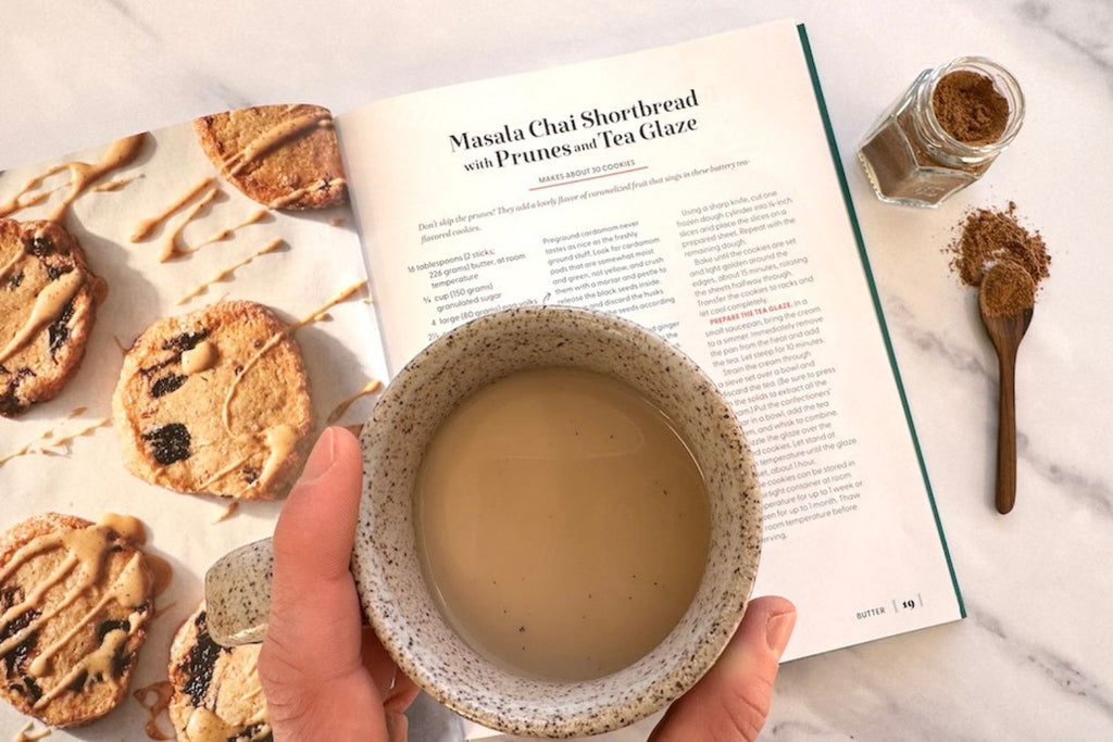 Hand holding a warm cup of Chai Masala tea over a cookbooks showing a photo of the chai tea cookies for the recipe included.