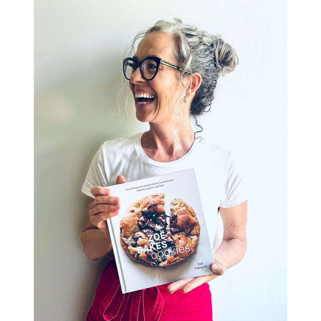 A smiling individual with curly hair and glasses holds a cookbook titled "Zoë Bakes Cookies: Everything You Need to Know to Make Your Favorite Cookies and Bars" by Zoë François. Dressed in a white shirt and red apron, they stand against a simple white background, eager to share delicious cookie recipes. The book is associated with the brand Gneiss Spice.