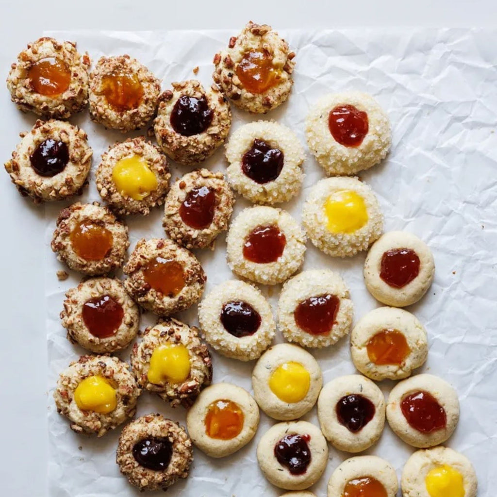 A delightful assortment of cookies from *Zoë Bakes Cookies: Everything You Need to Know to Make Your Favorite Cookies and Bars* by Gneiss Spice, artfully arranged on parchment paper, is adorned with vibrant jams in dark purple, yellow, red, and orange shades. These Zoë François creations are either enhanced with chopped nuts or left delectably plain—ideal for any cookie recipe collection.