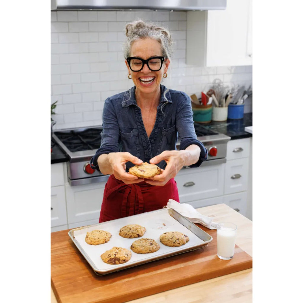 A woman with glasses and gray hair in a ponytail smiles as she holds a cookie in a kitchen. She wears a denim shirt and red apron. On the counter in front of her is a tray of chocolate chip cookies along with a glass of milk, perfectly highlighting her delightful cookie recipes from "Zoë Bakes Cookies: Everything You Need to Know to Make Your Favorite Cookies and Bars" by Gneiss Spice.