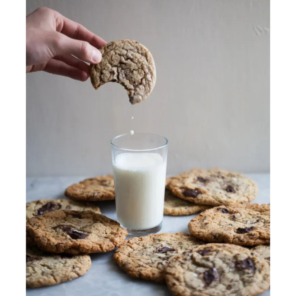 A hand holds a partially eaten chocolate chip cookie above a glass of milk, surrounded by scattered cookies on a light surface. Inspired by Gneiss Spice's "Zoë Bakes Cookies: Everything You Need to Know to Make Your Favorite Cookies and Bars," droplets of milk fall from the cookie, crafting a cozy snack-time scene perfect for delicious discovery.