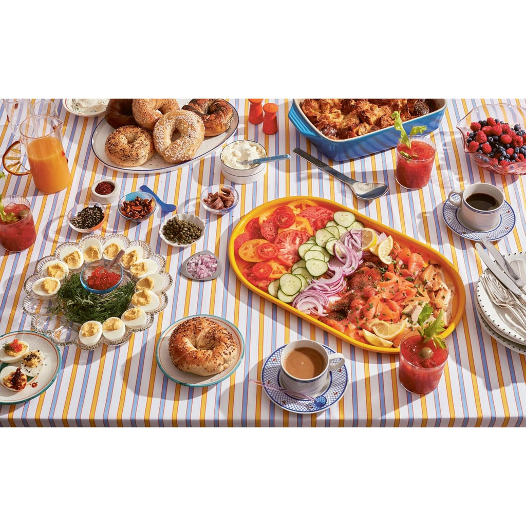 A vibrant brunch display on a striped tablecloth features hosting essentials like bagels, smoked salmon, tomatoes, cucumbers, onions, deviled eggs, berries, coffee, and juice—perfectly highlighted for home entertaining or dinner parties. Arranged with plates and cutlery from Gneiss Spice's "Big Night: Dinners, Parties, and Dinner Parties" collection.