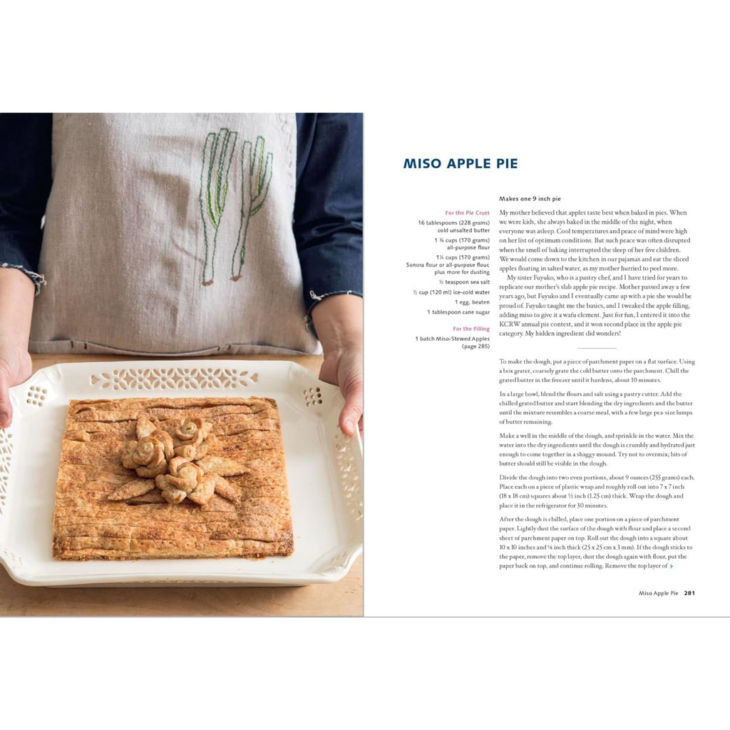 A person wearing a cactus-embroidered apron holds a rectangular pie on an ornate white tray, showcasing the essence of Japanese-inspired fusion cuisine. The pie is adorned with decorative pastry cutouts, while to the right, "Wafu Cooking: Everyday Recipes with Japanese Style" by Gneiss Spice invites adventurous home cooks with its intriguing ingredients and instructions for making the Miso Apple Pie.