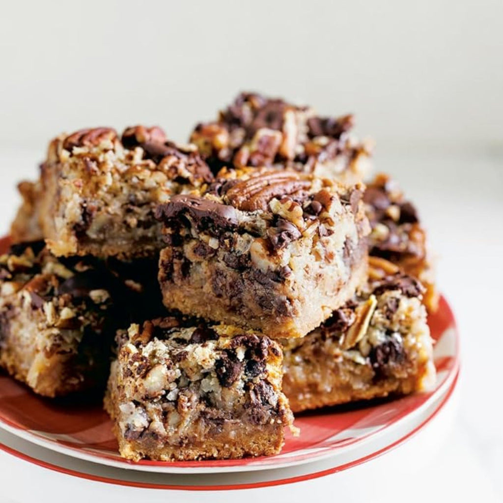 A stack of chocolate pecan squares, evoking the delicious creations found in "Zoë Bakes Cookies: Everything You Need to Know to Make Your Favorite Cookies and Bars" by Gneiss Spice, sits on a red and white striped plate. The squares feature a crumbly, nutty top layer adorned with visible chocolate chunks and pecans. The soft, neutral background perfectly complements Zoë François's delightful cookie recipes.