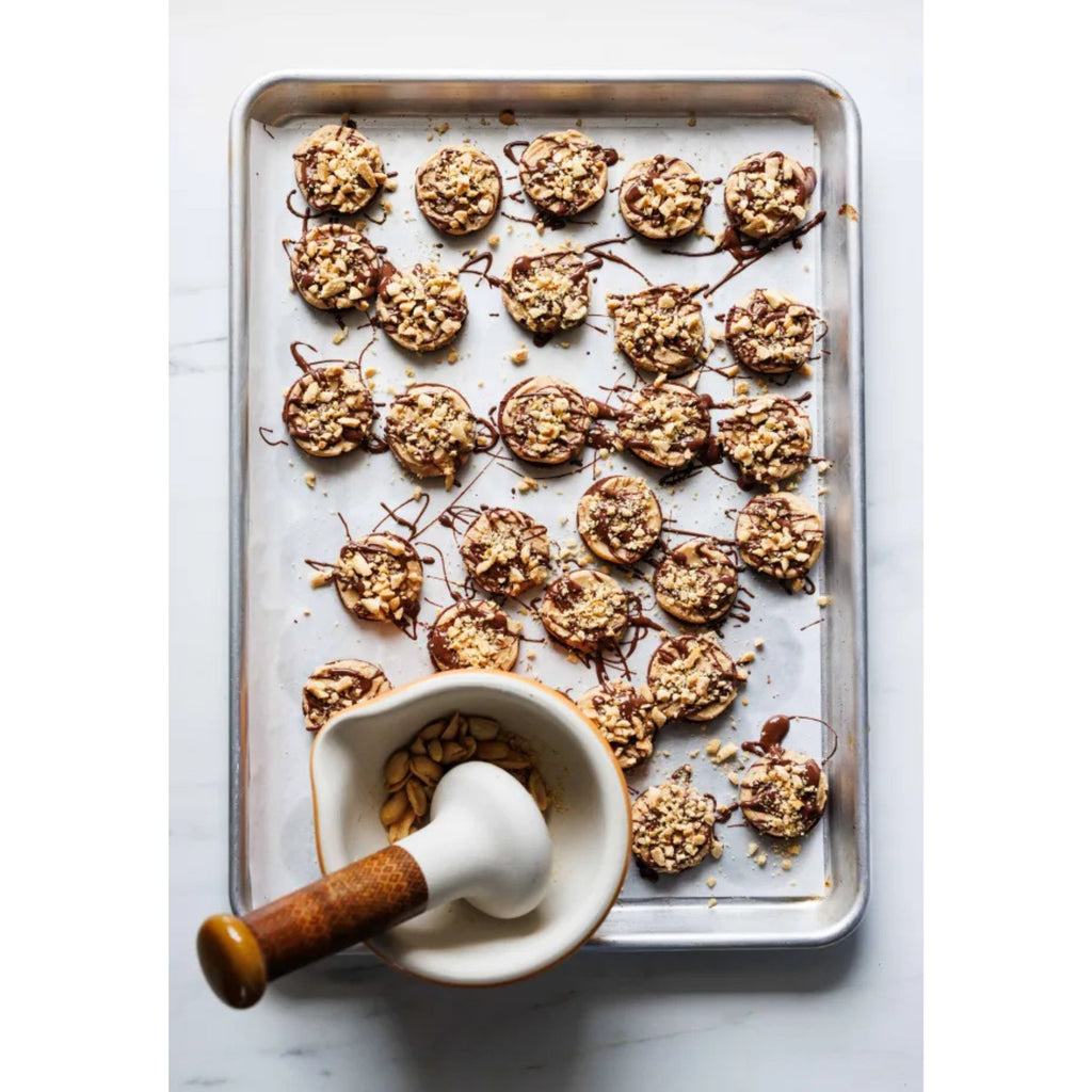 Displayed on a baking sheet are 24 round cookies from the book "Zoë Bakes Cookies: Everything You Need to Know to Make Your Favorite Cookies and Bars," topped with chopped nuts and drizzled with chocolate. In the foreground, there's a mortar and pestle from Gneiss Spice filled with nuts, capturing the essence of Zoë's delightful cookie recipes.