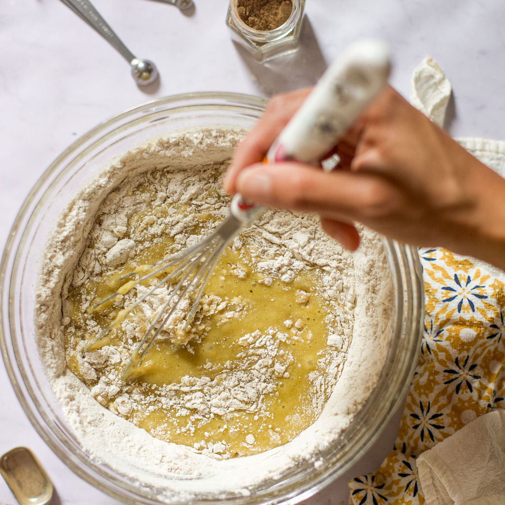 A person is whisking a mixture of flour and eggs in a glass bowl on a marble counter. Nearby, magnetic jars from the Gneiss Spice Baker's Bundle Holiday Gift Set, filled with organic baking spices, are arranged alongside measuring spoons and a towel with a floral pattern.