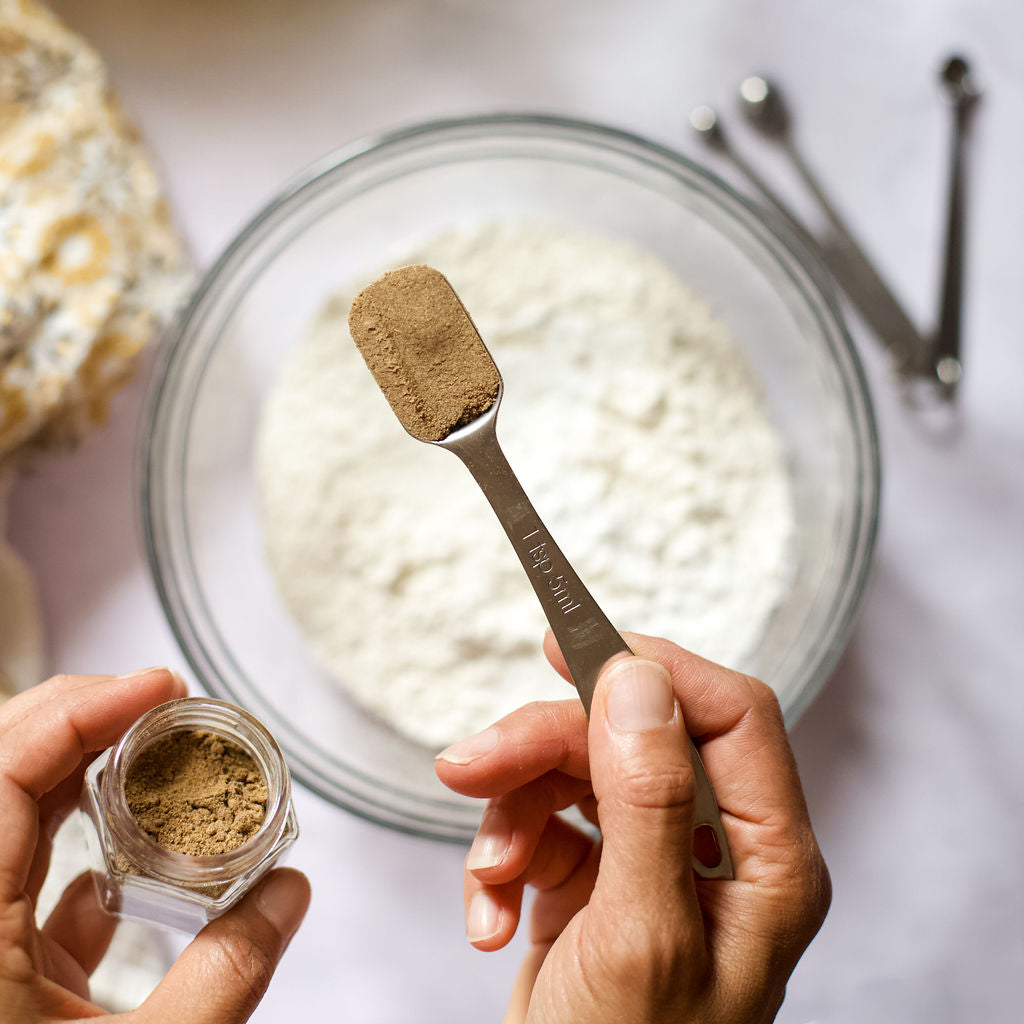 Close-up of a person measuring organic ground cinnamon with a teaspoon from a small, magnetic jar. Nearby, a glass bowl filled with flour rests on a white surface, accompanied by metal measuring spoons and a floral-patterned cloth—a delightful scene featuring the Gneiss Spice Baker's Bundle, an artisanal holiday gift set perfect for baking enthusiasts.