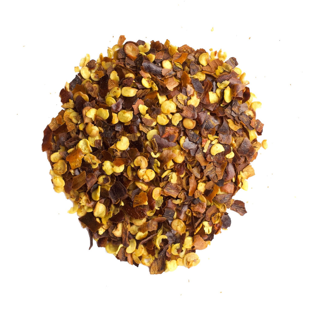 A pile of Gneiss Spice Red Pepper (Chili Flakes) on a white background showcases a blend of vibrant red and yellowish seeds, interspersed with small pieces of dried chili skin.