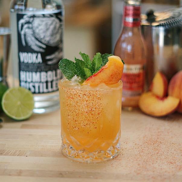 A refreshing cocktail in a glass garnished with a peach slice and mint leaves features a subtle kick from Gneiss Spice's Chili Fruit Salt Sprinkle & Drink Rim. Fresh peach slices and lime rest on the wooden table beside a bottle of vodka, with Zero-Waste Spice Refills subtly visible in the background.