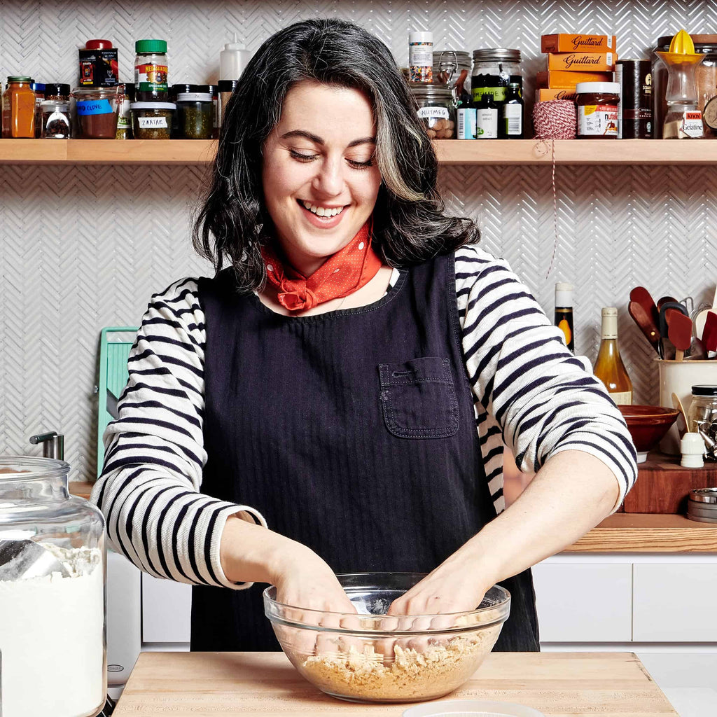 A smiling woman with long dark hair, wearing a striped shirt, black apron, and red scarf, mixes ingredients in a bowl. Surrounded by Gneiss Spice jars on shelves, she embodies the Dessert Person spirit from "Dessert Person: Recipes and Guidance for Baking with Confidence," crafting a delightful recipe.