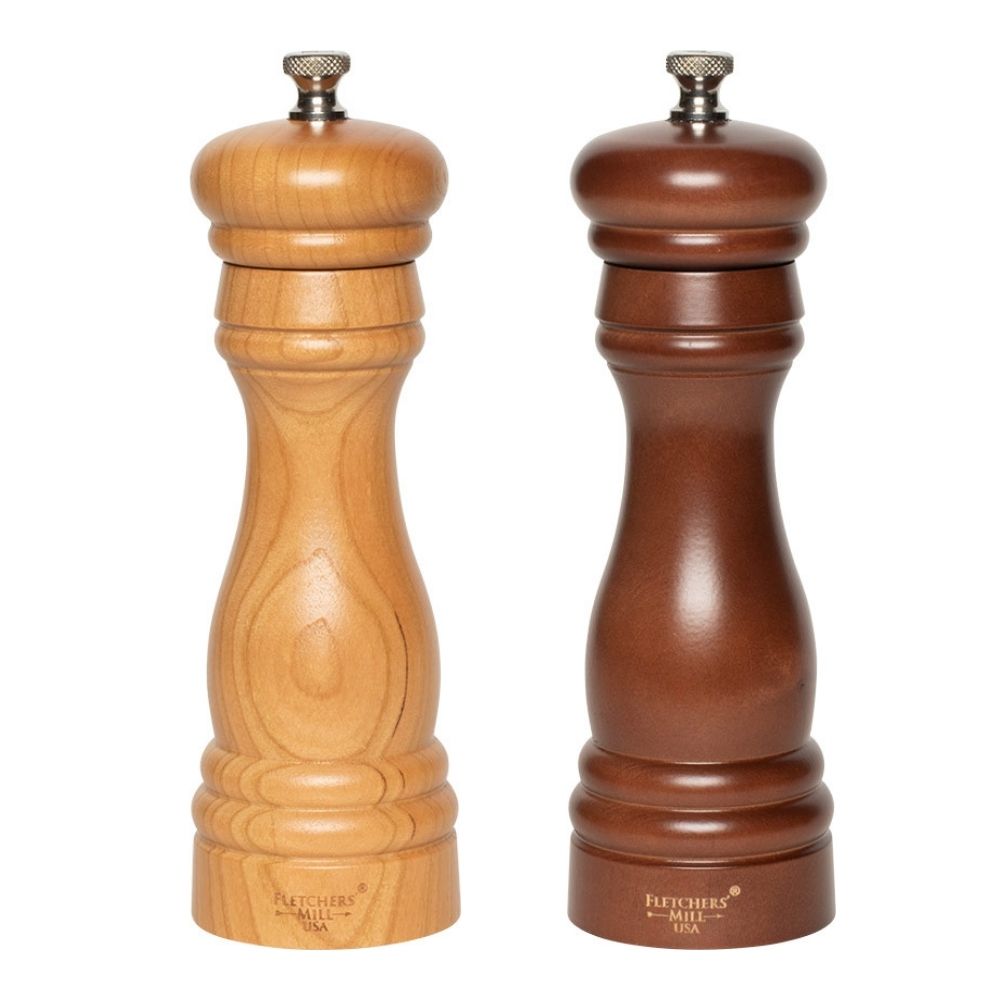 Two wooden grinders from the Gneiss Spice Grinder Gift Set are positioned next to each other. The left one features a light brown finish, while the right one is dark brown. Both are made from sustainably-harvested wood and boast a classic design, complete with metallic knobs on top and adjustable coarseness settings. The base of each grinder is engraved with "Fletcher's Mill USA.