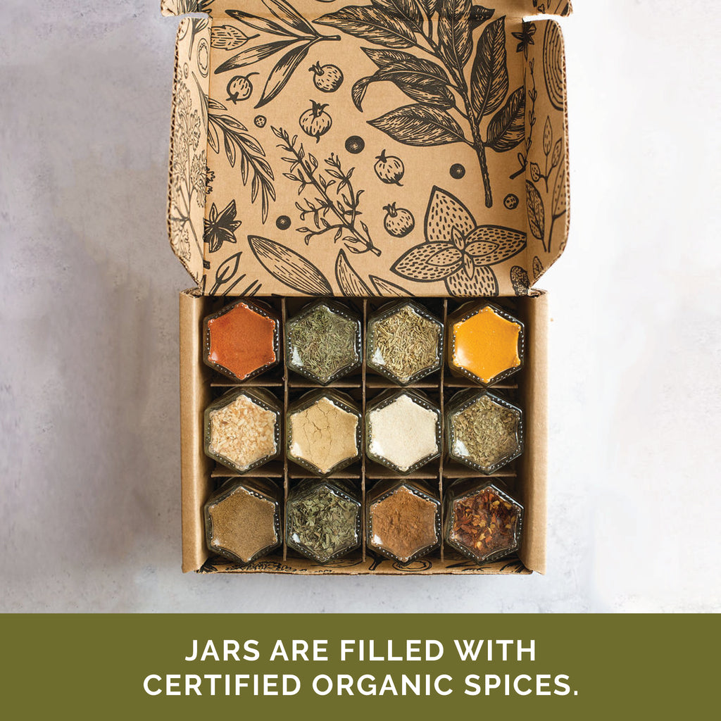 A set of twelve magnetic jars from Gneiss Spice, each with a hexagonal lid and filled with certified organic spices ranging in colors from orange to green to white, lies open. Inside the cardboard box, botanical illustrations are displayed.