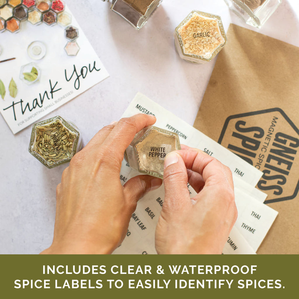 Hands holding a hexagonal spice jar labeled "White Pepper" from the Gneiss Spice BASICS set. Nearby are other organic spice jars, a "Thank You" note, and a sheet featuring clear and waterproof spice labels. A brown bag with "Chesapeake Spice" branding is in the background.