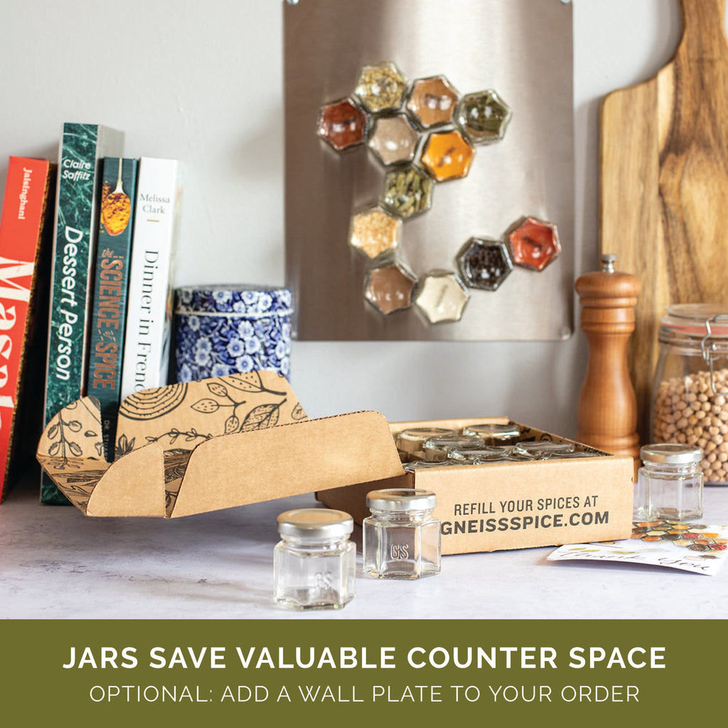 In a cozy kitchen setting, a wall display of Gneiss Spice's BASICS collection showcases hexagonal magnetic jars filled with organic spices. A cardboard box containing additional spice jars is positioned on the counter next to cookbooks, a pepper grinder, and a plant. The text highlights that these jars help save valuable counter space and suggests adding an optional wall plate to your order.