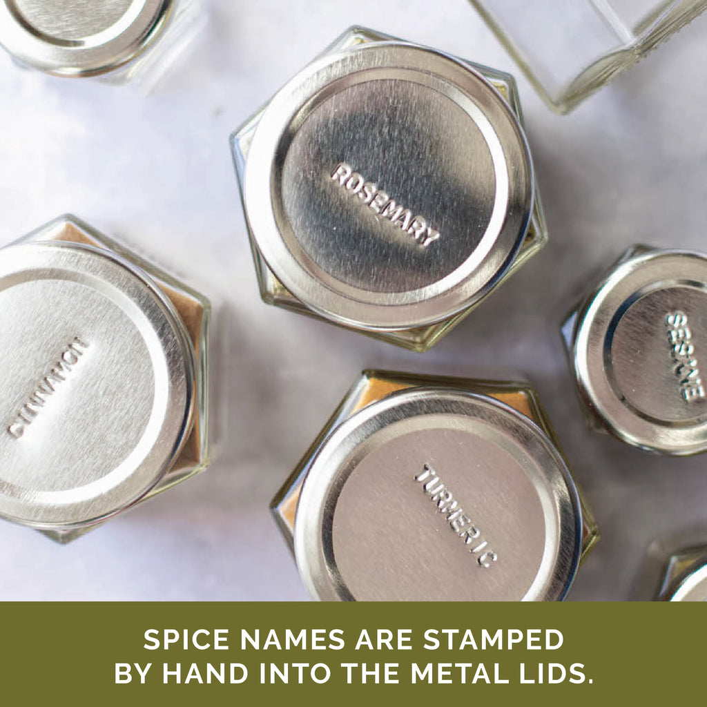 Twelve hexagonal magnetic jars filled with organic spices from Gneiss Spice feature metal lids, each hand-stamped with the name of a spice like rosemary, cinnamon, or turmeric. A green strip at the bottom reads, "Organic spice names are stamped by hand into the metal lids.