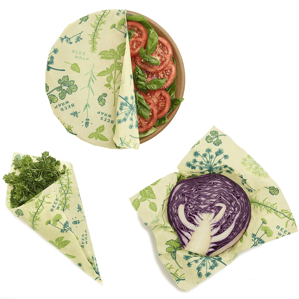 Gneiss Spice's Beeswax Wrap 3-Pack, made from organic cotton with various patterns, covers a bowl of sliced tomatoes and wraps fresh parsley and half a purple cabbage for sustainable food storage.