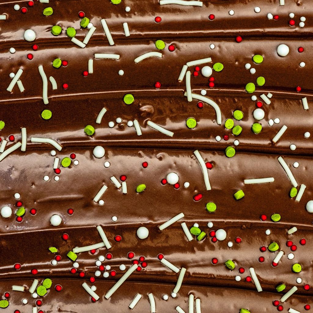 Close-up of a chocolate surface adorned with Gneiss Spice's "Jingle Pop" holiday sprinkles in festive green, red, and white colors. The sprinkles feature small balls and thin strands scattered across the smooth, rich brown chocolate.