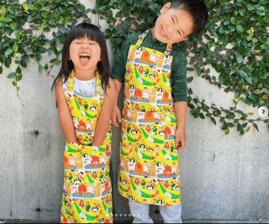 Two joyful children in Gneiss Spice Limited Edition Aprons + Tea Towels stand by a leafy wall. The girl laughs with hands in pockets, and the boy grins with his head tilted. These aprons boast vibrant prints inspired by Joelle Avellino illustrations.