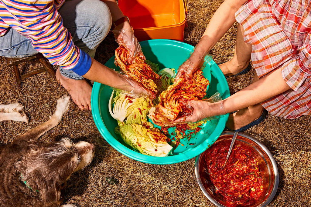 Two people, a dog nearby, blend cabbage with red seasoning in a large green bowl. A metal bowl of extra seasoning sits beside them on dry grass. One wears gloves; the other, a striped shirt—capturing techniques from "Korean American: Food That Tastes Like Home" by Gneiss Spice.