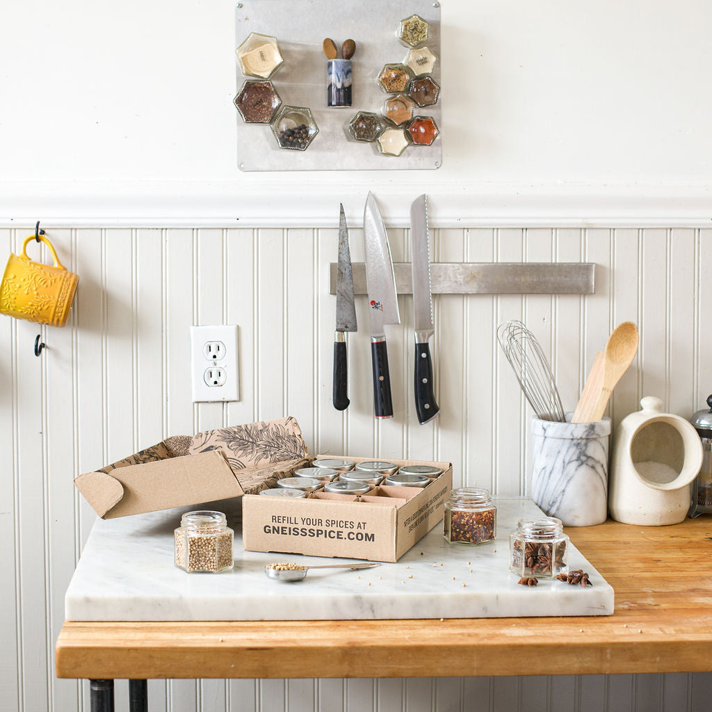 A kitchen scene with a marble-topped wooden table, showcasing a 'FOODIE' GIFT BUNDLE from Gneiss Spice, which includes empty magnetic jars and a gift certificate for organic refills. The scene also features a set of knives on a magnetic strip, a white jar containing utensils, and a marble mortar and pestle. Hexagonal magnetic spice jars are neatly mounted on the wall.