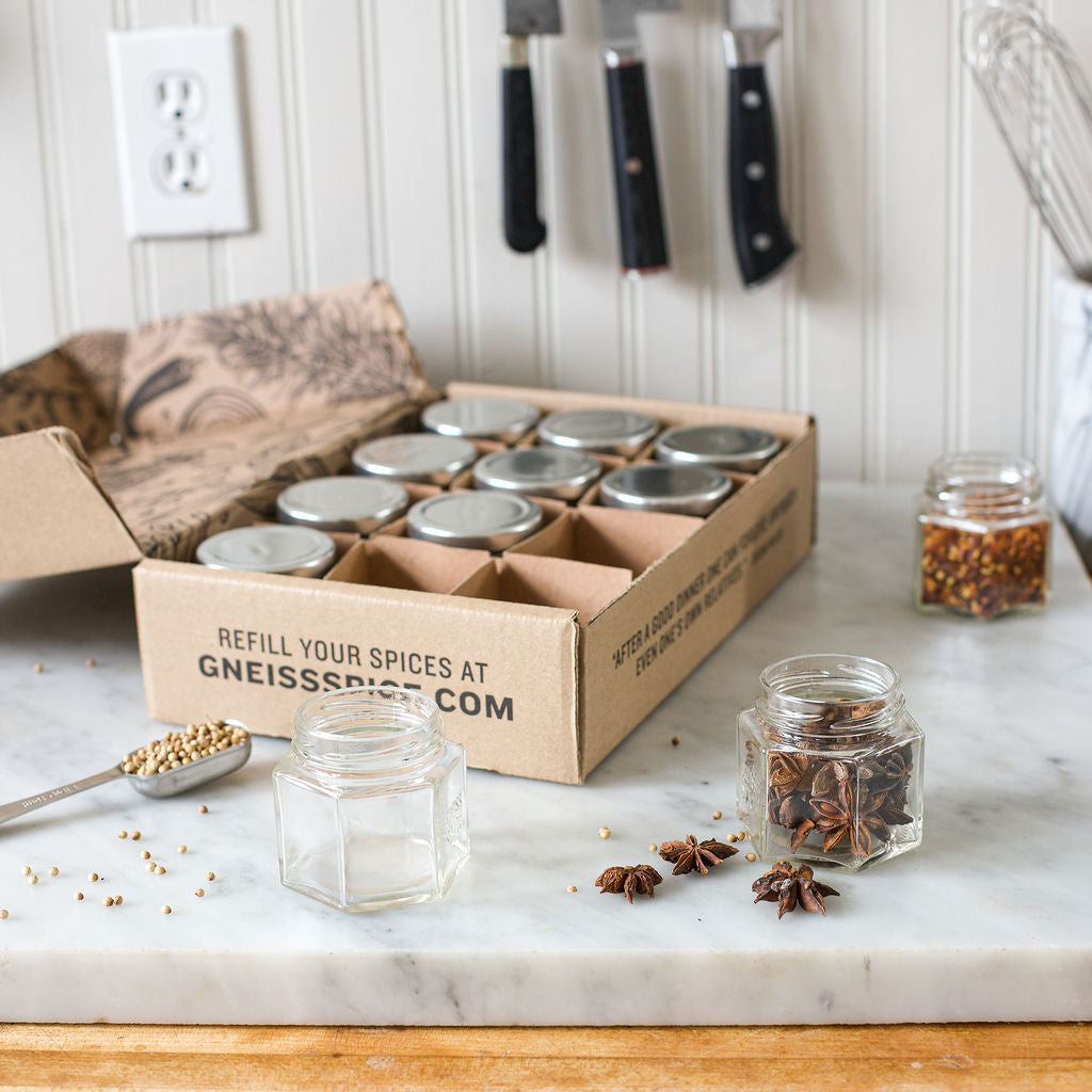 A kitchen countertop features the 'FOODIE' GIFT BUNDLE by Gneiss Spice, showcasing a box of empty magnetic jars ready for filling with a gift certificate for organic refills. Knives are neatly arranged on a wall rack nearby, while an open jar holds star anise and chili flakes amidst scattered seeds.