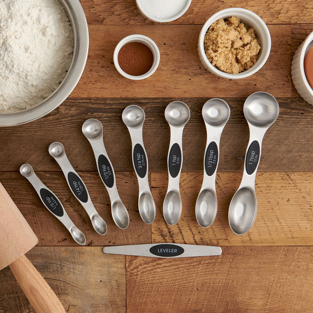 A set of Gneiss Spice's Mrs Anderson's Magnetic Measuring Spoons, labeled from 1/8 tsp to 1 tbsp and featuring a leveler, is neatly arranged on a wooden surface. Nearby, you'll find bowls of flour, sugar, and cinnamon, accompanied by an egg and a rolling pin.