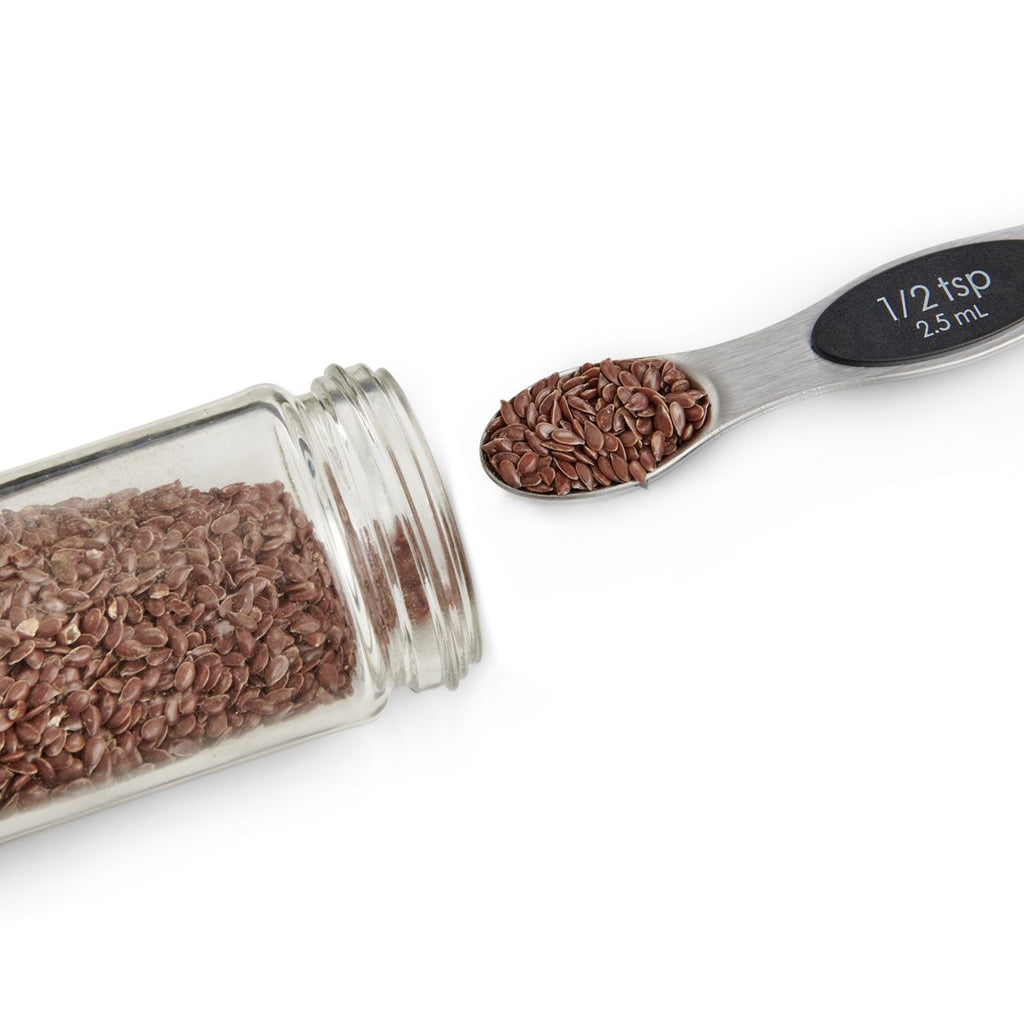 A glass jar of flaxseeds is tipped over beside a magnetic measuring spoon from Mrs Anderson's, branded by Gneiss Spice, labeled "1/2 tsp" and filled with seeds, set against a pristine white background.