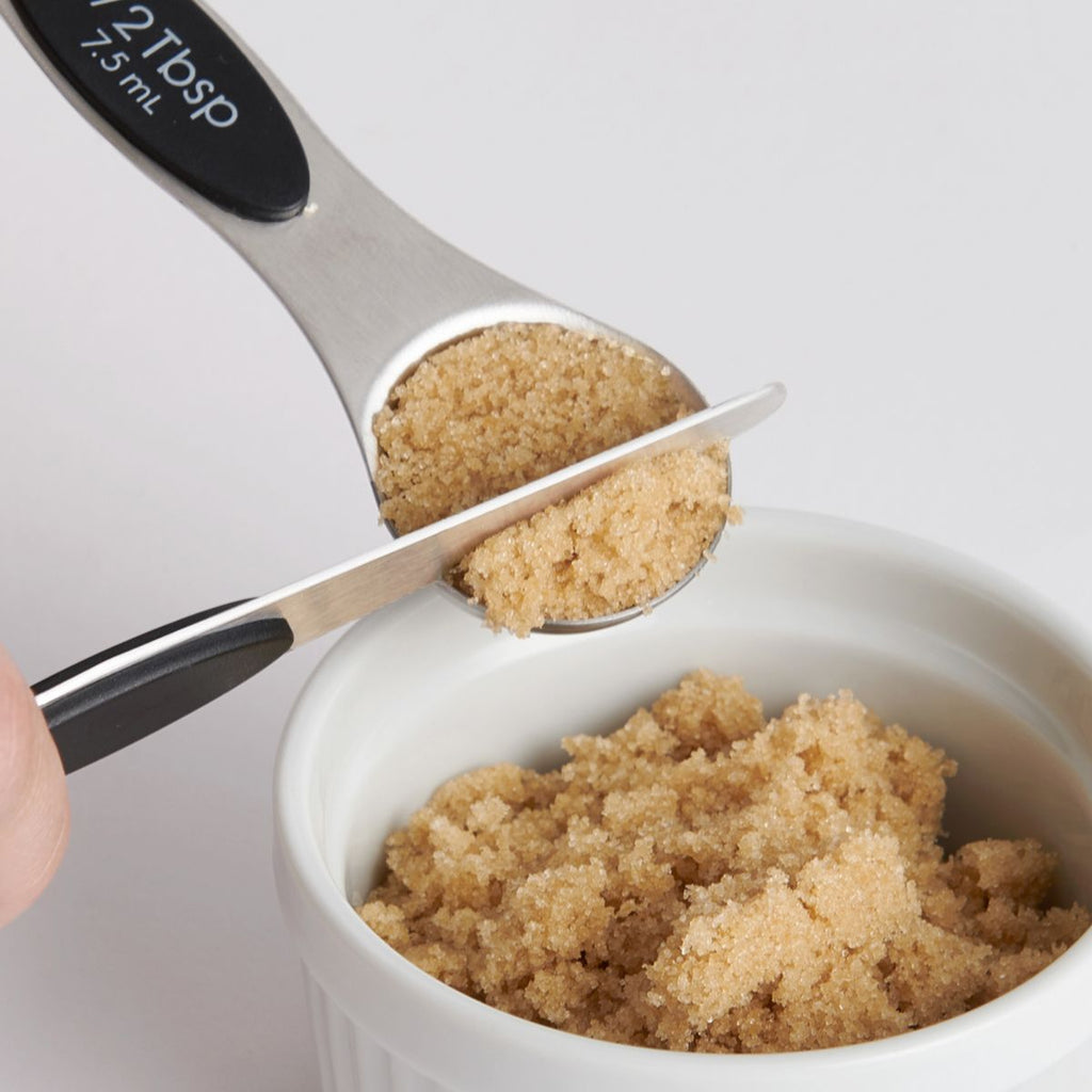 A close-up reveals a tablespoon from the Gneiss Spice Mrs Anderson's Magnetic Measuring Spoons set being used to level brown sugar in a white ramekin. The person adeptly scrapes off the excess with a flat edge. The stainless steel utensil is engraved with "2 Tbsp" and "25 mL.