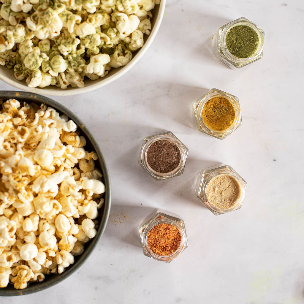 Two bowls of gluten-free popcorn, one garnished with vibrant green seasoning and the other left plain, are beautifully displayed on a marble surface. Nearby, five small jars feature the hand-blended seasonings from Gneiss Spice's Popcorn Spice Kit — Your Choice of 3 Seasonings, offering an assortment of organic blends to suit any palate.