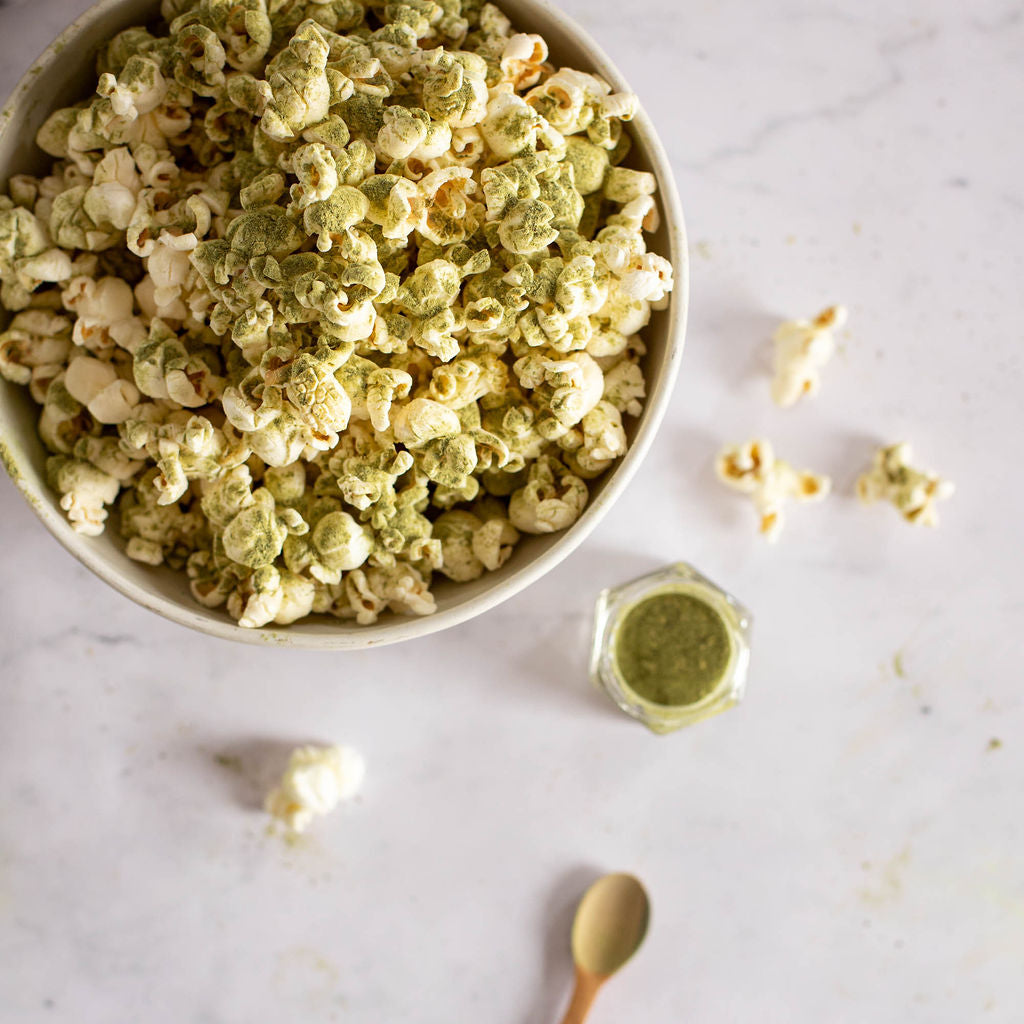 A bowl of popcorn, crafted for vegans and topped with green matcha powder, rests on a marble surface. Beside it, a small jar from the Gneiss Spice Popcorn Spice Kit — featuring 7 organic seasonings perfect for movie night — is accompanied by a wooden spoon. A few pieces of gluten-free popcorn are scattered nearby.