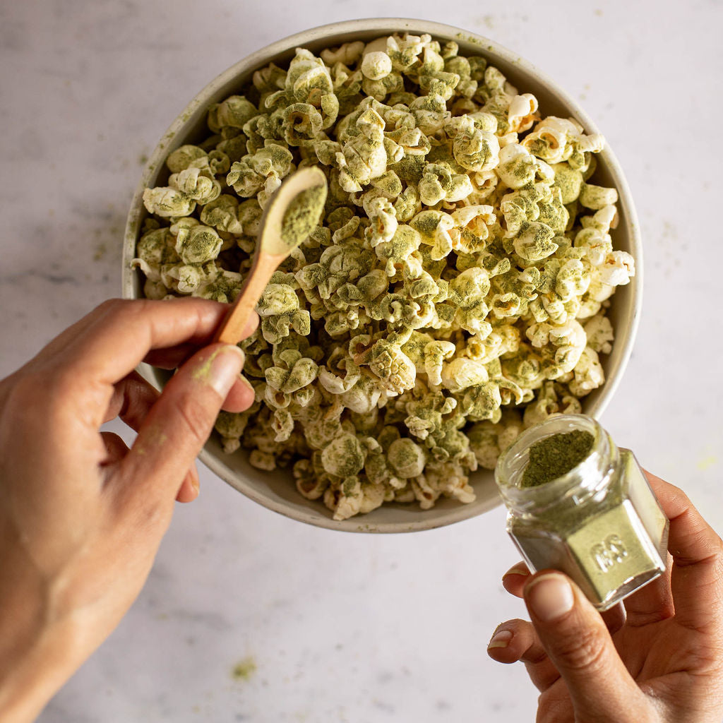 A person uses a small wooden spoon to sprinkle matcha powder from the Popcorn Spice Kit by Gneiss Spice over a bowl of popcorn. The kit, which includes an assortment of 12 organic seasonings for a gluten-free movie night, features a small jar of matcha visible in one hand. The scene is elegantly set on a marble surface.