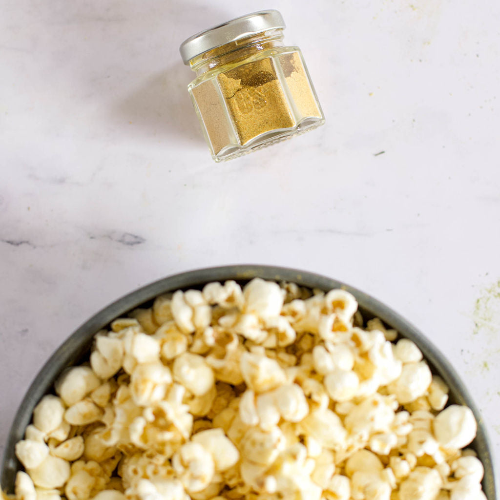 A bowl of popcorn blends sits on a white marble surface, made enticingly with the Popcorn Spice Kit from Gneiss Spice. Nearby, a small jar from this kit containing one of the seven organic seasonings is placed at an angle, emphasizing its vegan-friendly appeal for enhancing your movie night treats.