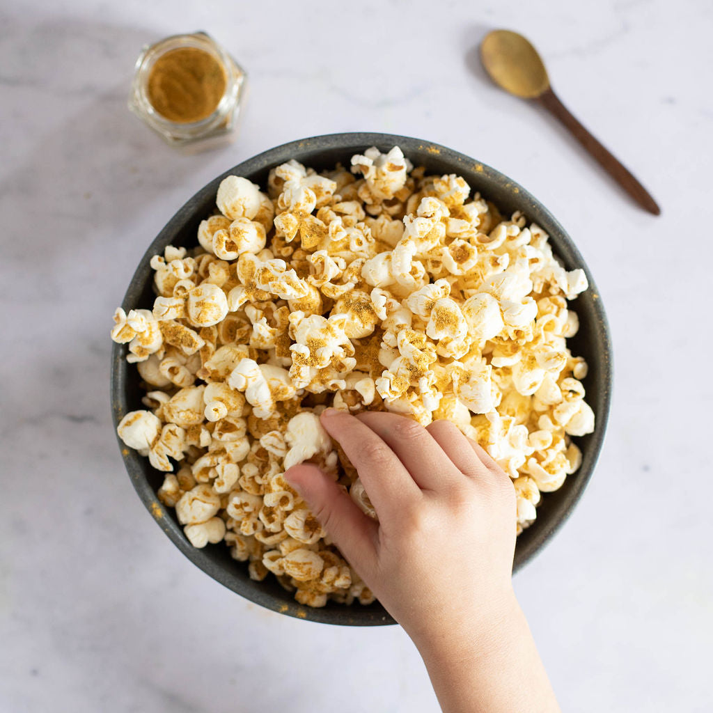 A hand reaches into a bowl of popcorn on a marble surface. Nearby, a Gneiss Spice eco-friendly jar from the Popcorn Spice Kit and a spoon sit ready to add flavor. The organic seasonings enhance the lightly seasoned treat.