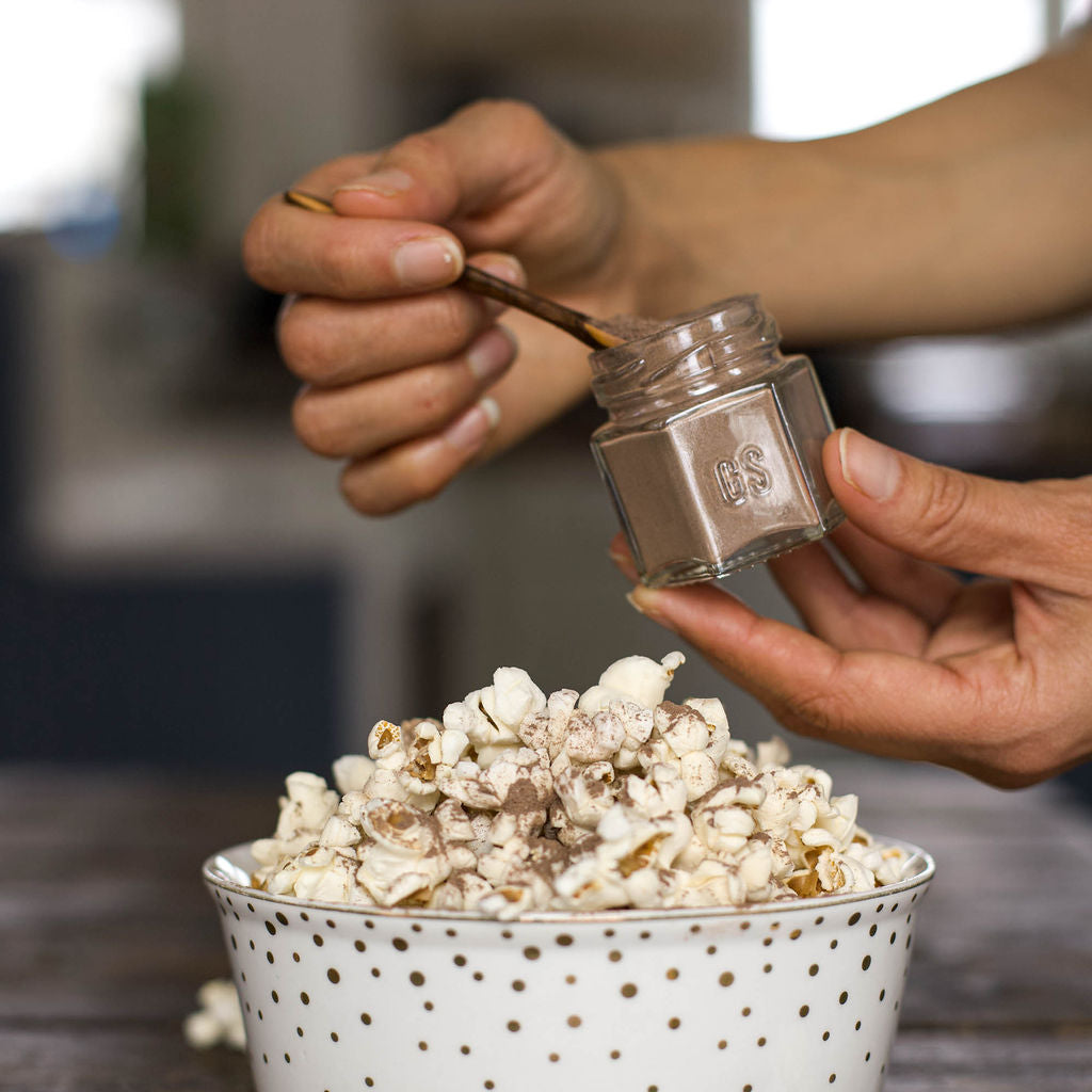 A person sprinkles organic seasoning from a small jar onto vegan-friendly popcorn in a white bowl with gold dots, using the Gneiss Spice (Sale) Popcorn Spice Kit — 7 Organic Seasonings for Movie Night. The background is softly blurred.