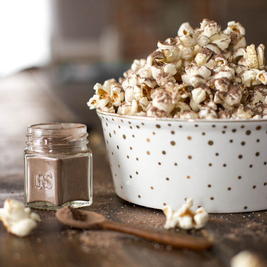 A bowl of vegan-friendly popcorn, seasoned with Gneiss Spice's (Sale) Popcorn Spice Kit — featuring 7 Organic Seasonings for Movie Night, sits on a wooden table. Nearby, there is a glass jar of cocoa powder and a wooden spoon with some spilled powder. The white bowl, adorned with brown polka dots, adds charm to the gluten-free snack setting.