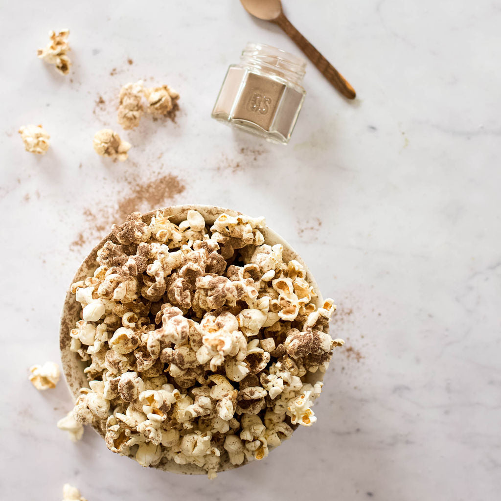 A bowl of gluten-free popcorn, lightly dusted with cinnamon, rests on a marble surface. Nearby, there's a small jar of powdered cinnamon accompanied by a teaspoon and scattered pieces from the Gneiss Spice Popcorn Spice Kit — 12 Organic Seasonings for Movie Night.