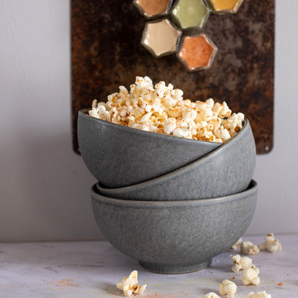 A cozy vegan-friendly snack setup is captured with two stacked gray bowls filled with gluten-free popcorn, garnished with a pinch from the (Sale) Popcorn Spice Kit — 7 Organic Seasonings for Movie Night by Gneiss Spice, set against a backdrop of spice hexagons. A few pieces are playfully scattered on the white surface.