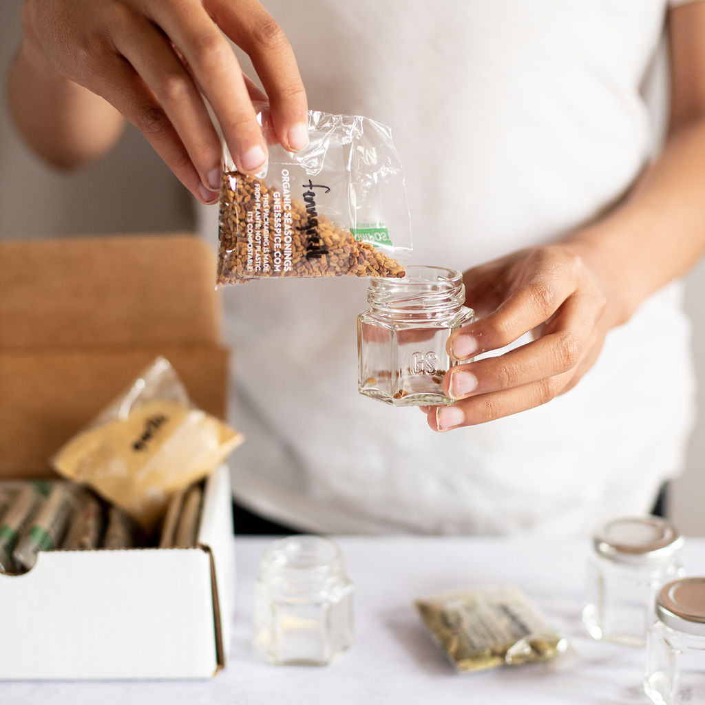 A person in a white shirt is transferring organic seasonings from a small bag into one of the magnetic jars from the Gneiss Spice 'FOODIE' Gift Bundle. Several other bags and empty jars are neatly arranged on the white surface nearby, with part of a cardboard box visible in the background.