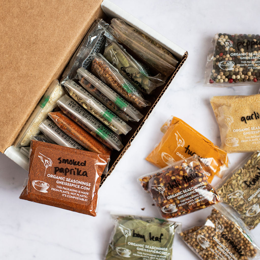 A collection of seasoning packets, such as smoked paprika and organic blends, are artistically arranged around and inside a cardboard box. Some of these feature personalized spice labels, while additional packets containing seeds and herbs are splayed across a marble surface. This is part of the 'FOODIE' GIFT BUNDLE by Gneiss Spice, which includes empty magnetic jars and a gift certificate for organic refills.