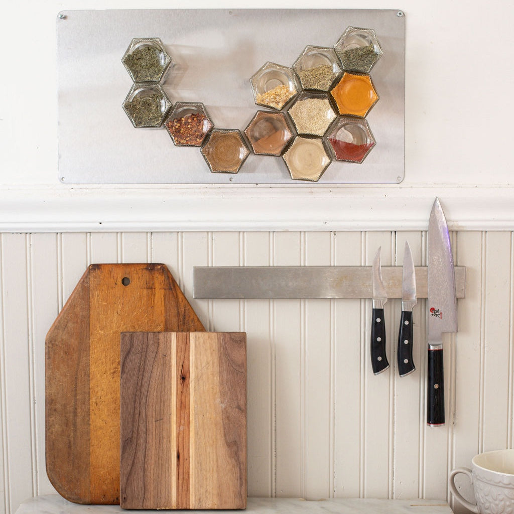 In the kitchen scene, Gneiss Spice's BASICS magnetic jars filled with organic spices are neatly arranged on a wall-mounted metal board. Nearby, a knife magnet holds three knives, while two wooden cutting boards lean against the white-paneled wall.