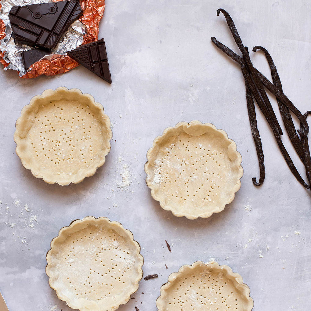 On a gray surface, four unbaked tart crusts are arranged alongside pieces of fair trade dark chocolate on an orange foil wrapper in the top left corner, while Gneiss Spice's certified organic and fair trade Gourmet Vanilla Pods from Madagascar rest on the right, creating an exquisite baking scene.