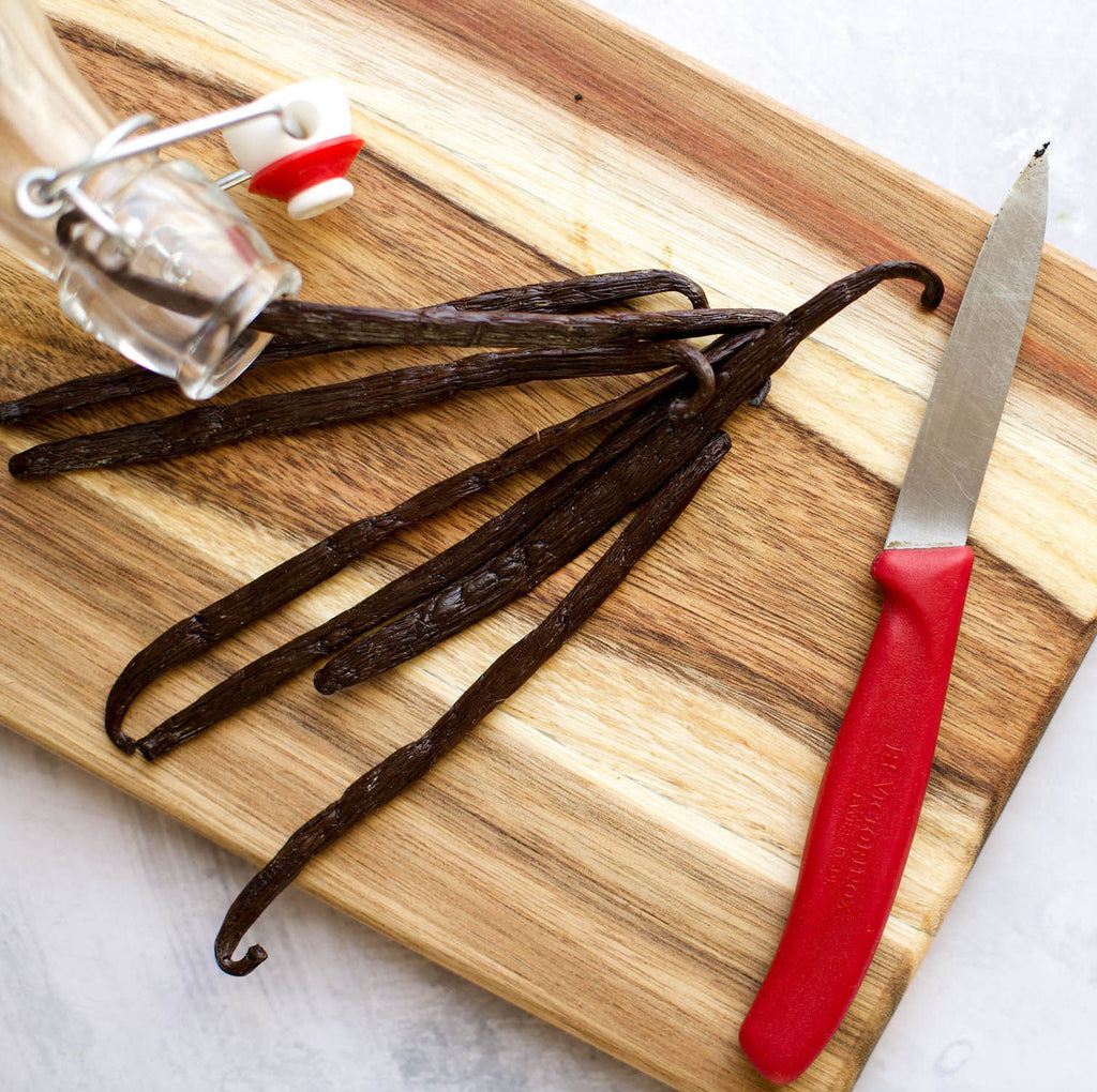 A wooden cutting board displays Gneiss Spice's Gourmet Vanilla Pods - Certified Organic & Fair Trade from Madagascar, alongside an empty glass bottle with a swing top and a knife with a red handle. Placed on a light kitchen surface, this elegant arrangement inspires culinary creativity.