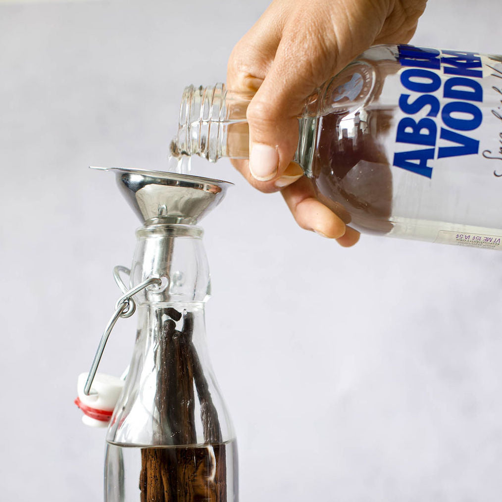 A hand pours vodka from an Absolut bottle into a glass bottle with the aid of a metal funnel. This glass bottle is elegantly adorned with Gneiss Spice Gourmet Vanilla Pods - Certified Organic & Fair Trade from Madagascar, and has a striking red and white stopper. The setting is minimalistic, highlighted by a simple light background.