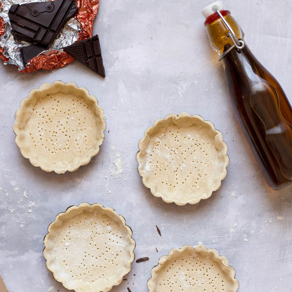 Four unbaked pie crusts rest in round tins on a gray surface. Beside them, an unwrapped chocolate bar sits on foil alongside a glass bottle of dark liquid with a stopper—part of the Baker's Bundle by Gneiss Spice, a delightful holiday gift set with baking spices, tools, and kit to make homemade vanilla extract.