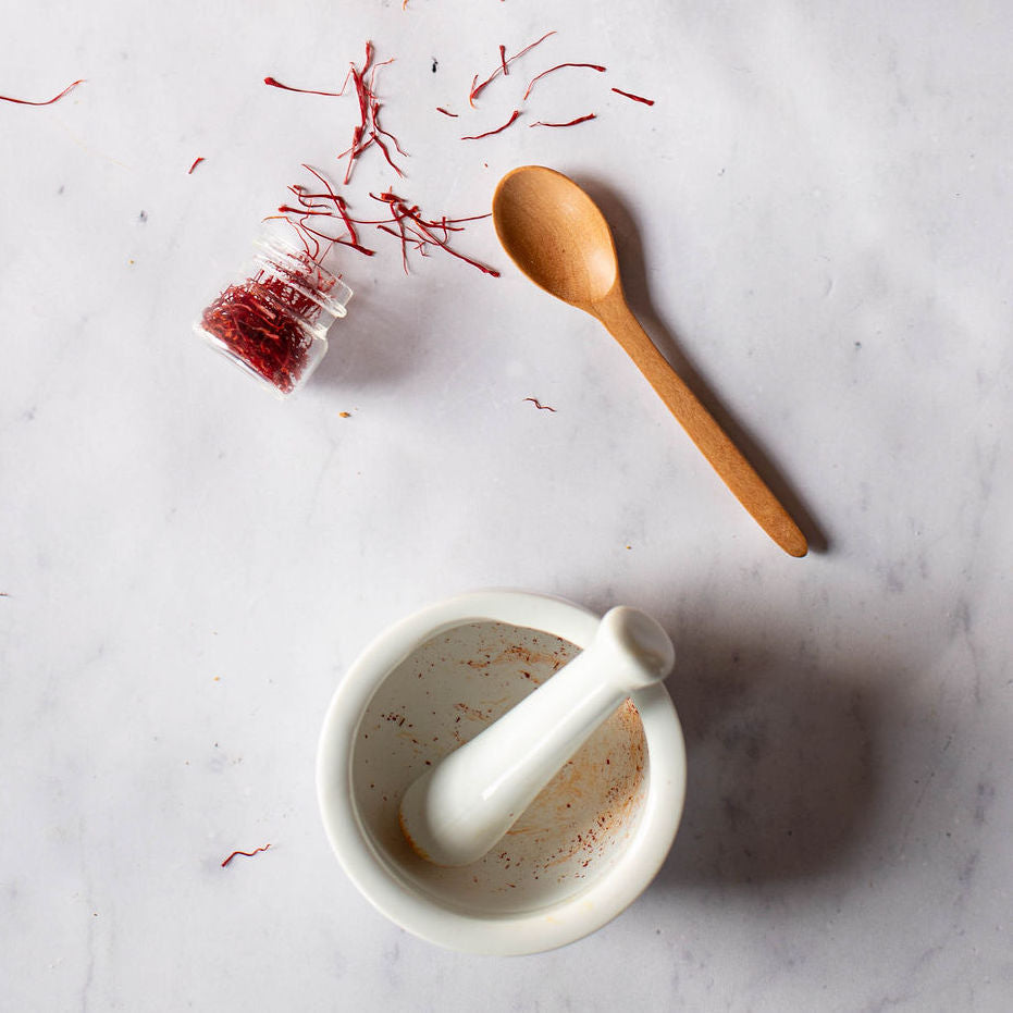 A small jar of Gneiss Spice saffron spills across a marble surface beside a wooden spoon, evoking the rich flavors of Indian cooking. A white mortar and pestle with saffron residue hints at a recipe in progress, weaving aromatic magic into Middle Eastern dishes.