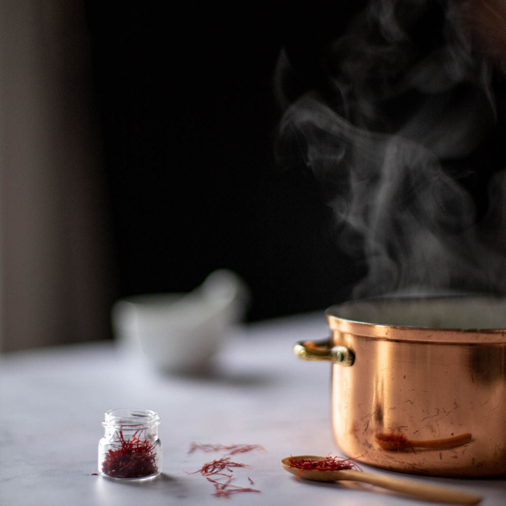 A steaming copper pot sits on a table beside a small glass jar of Gneiss Spice's Saffron. Infusing a touch of Indian cooking, a wooden spoon featuring saffron and blurred background adds a cozy kitchen ambiance.