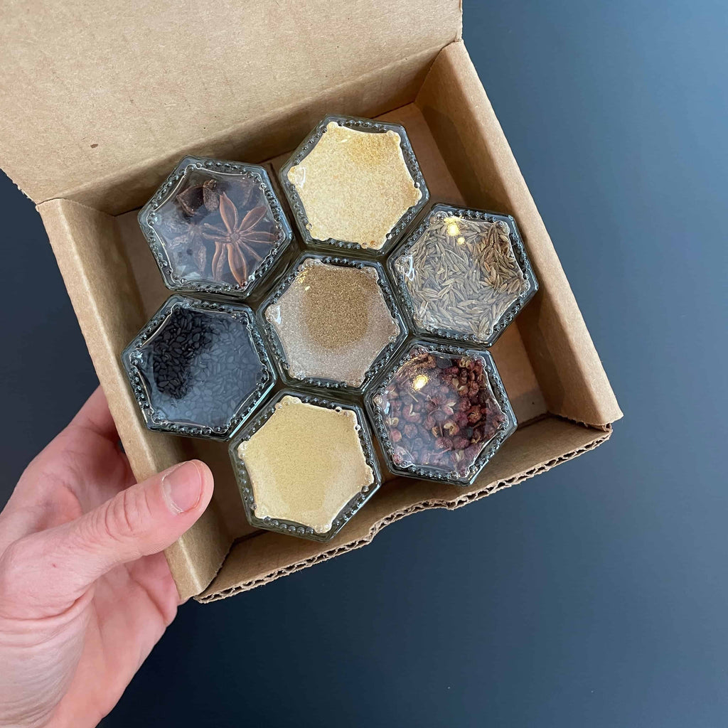 A hand holds an open cardboard box containing seven hexagonal glass jars with organic spices from the Sichuan Spice Kit by Gneiss Spice, showcasing black seeds, pale powders, and star anise. The spice kit is resting on a dark surface.