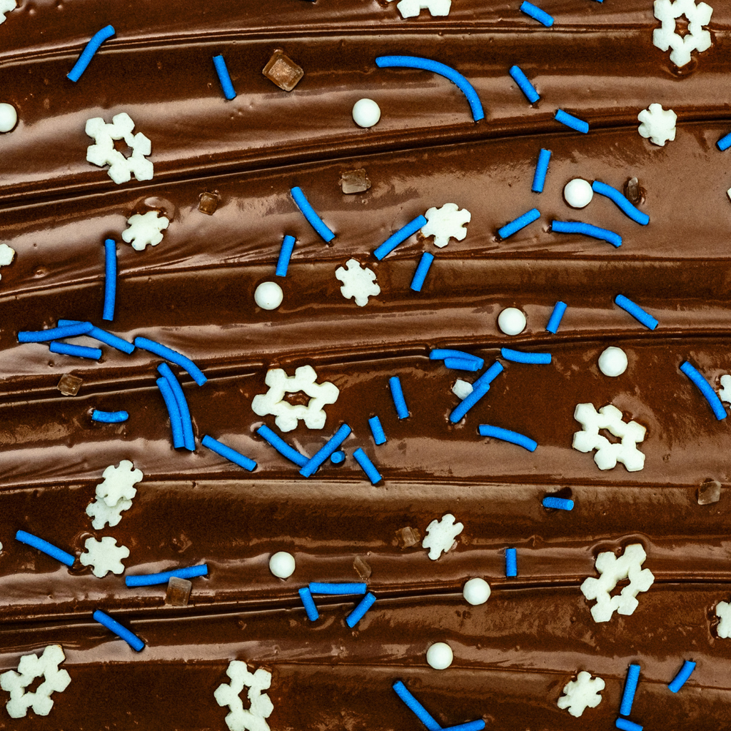 A close-up of Gneiss Spice's Sprinkles — All-Natural Holiday "Snow Fall" shows chocolate frosting adorned with blue and white winter sprinkles, featuring delicate long strands and intricate snowflakes. The frosting displays a smooth surface with small ridges, offering a textured look, all crafted with natural ingredients for a delightful finish.