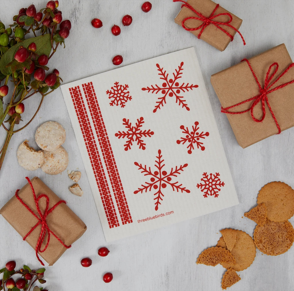 On a wooden surface sits a Swedish dishcloth adorned with red snowflake patterns and stripes. Surrounding it are eco-friendly gifts wrapped in brown paper and tied with red string, assorted cookies, and holly branches with red berries, all from Gneiss Spice's Holiday Promotion — Buy 3, Get 1 Free collection, creating a picturesque holiday scene.