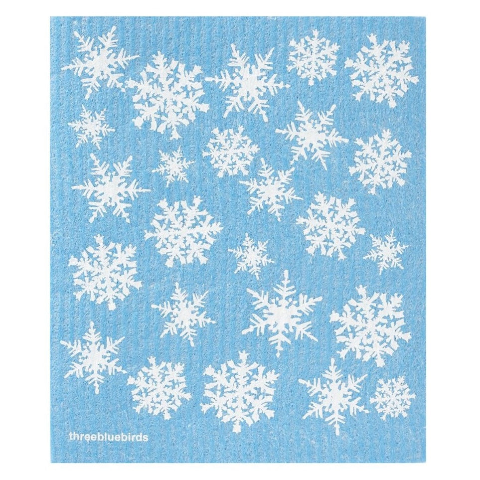 A Swedish dishcloth from Gneiss Spice, part of their "Holiday Promotion — Buy 3, Get 1 Free" offer, features a blue color with white snowflakes scattered across its surface. This eco-friendly cloth is reusable for sustainable cleaning and displays the text "threebluebirds" in the bottom left corner.